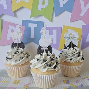 May include: Three cupcakes in white paper liners topped with white frosting and crushed cookie pieces. Each cupcake features a decorative topper: one with a duck in boots, one with a duck in a party hat, and one with a green gingham bow.