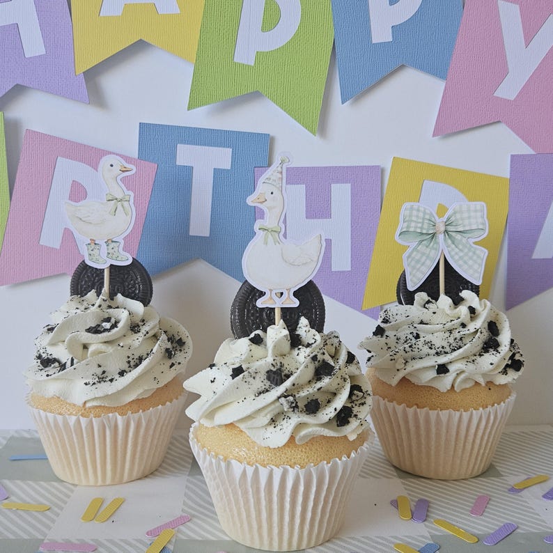 May include: Three cupcakes in white paper liners topped with white frosting and crushed cookie pieces. Each cupcake features a decorative topper: one with a duck in boots, one with a duck in a party hat, and one with a green gingham bow.