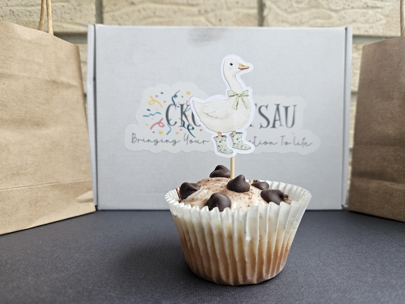 Puede incluir: Un cupcake con chispas de chocolate y un adorno de ganso. El ganso lleva botas de agua y un lazo. Una caja blanca con una pegatina y el texto "CK'SAU Bringing Your Vision to Life" en el fondo.