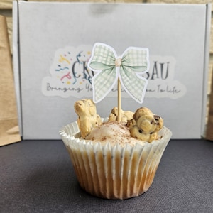 Puede incluir: Un cupcake decorado con un lazo de cuadros verde claro sobre un palito. El cupcake tiene un glaseado blanco con canela y galletas con forma de oso. En el fondo, una caja con el texto "CK SAU Bringing You From To Life".
