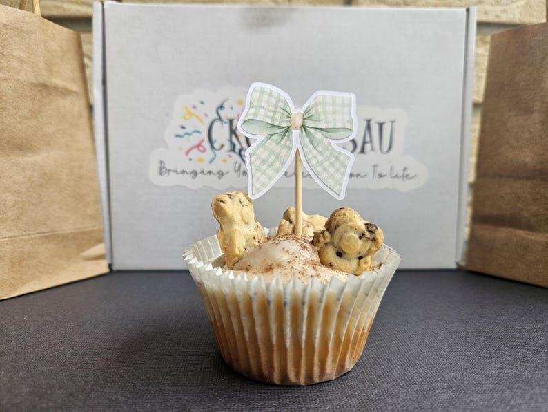 Puede incluir: Un cupcake decorado con un lazo de cuadros verde claro sobre un palito. El cupcake tiene un glaseado blanco con canela y galletas con forma de oso. En el fondo, una caja con el texto "CK SAU Bringing You From To Life".