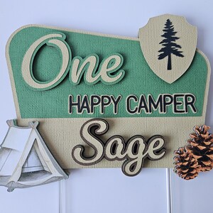 May include: A layered, decorative sign with the words "One Happy Camper Sage". The sign features a green and tan color scheme, a tent, a pine tree, and two pine cones. The sign is likely for a birthday or camping-themed event.