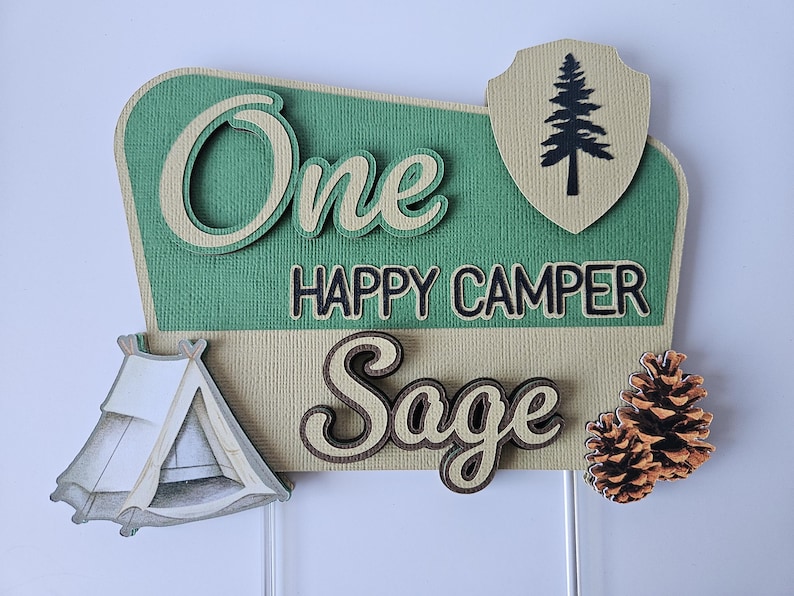 May include: A layered, decorative sign with the words "One Happy Camper Sage". The sign features a green and tan color scheme, a tent, a pine tree, and two pine cones. The sign is likely for a birthday or camping-themed event.