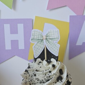 May include: A cupcake topper featuring a light green and white gingham patterned bow on a wooden stick, inserted into a cupcake with white frosting and dark cookie crumbles, set against a background of colorful paper birthday banner flags.