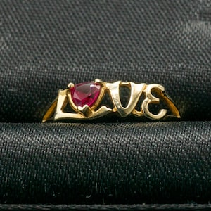 May include: A gold ring with the word "LOVE" spelled out in capital letters. The "O" is replaced by a heart-shaped, deep red gemstone. The ring is set against a black textured background.