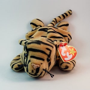May include: A brown and black striped stuffed tiger with a red and white Ty Beanie Baby tag. The tag has a white star and the words "Ty Beanie Baby" on it.