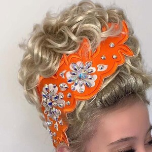 May include: A young girl with blonde curly hair wearing an orange and white hairpiece with rhinestones. The hairpiece is decorated with a floral design.