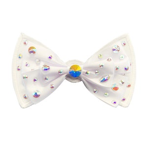 May include: A white bow adorned with iridescent rhinestones. The bow is crafted from a sheer material and features a prominent rhinestone at its centre. The edges are finished with white stitching. Suitable for hair or clothing.
