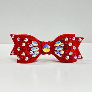 May include: A vibrant red bow decorated with clear and iridescent rhinestones. The bow is crafted from a soft, velvety fabric and has a central iridescent stone. This accessory is suitable for hair or clothing, and is approximately 10 cm wide.
