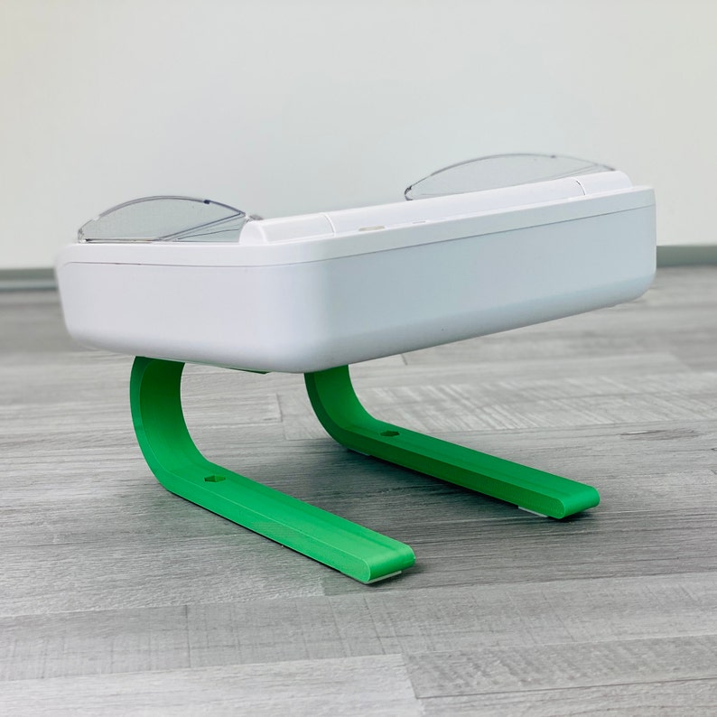 May include: White pet feeder with two clear plastic food bowls and a green base. The feeder has a modern design and is elevated on two curved legs.
