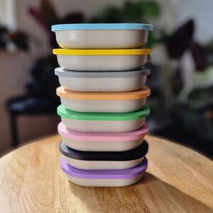May include: A stack of nine colorful round containers with a light blue lid on top. The containers are stacked in a rainbow pattern, starting with light blue, yellow, gray, beige, orange, green, pink, black, and purple.
