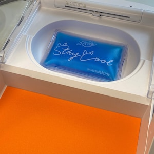 May include: A blue gel pack with the words "Stay Cool" in white script is inside a white compartment. The compartment is part of a larger white structure, with an orange surface visible below.