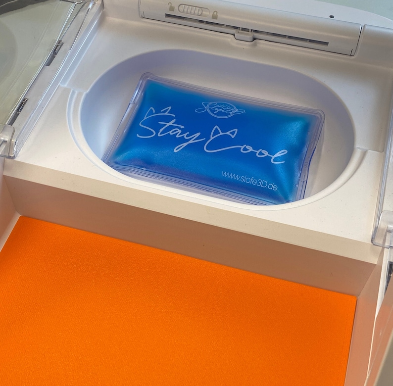 May include: A blue gel pack with the words "Stay Cool" in white script is inside a white compartment. The compartment is part of a larger white structure, with an orange surface visible below.