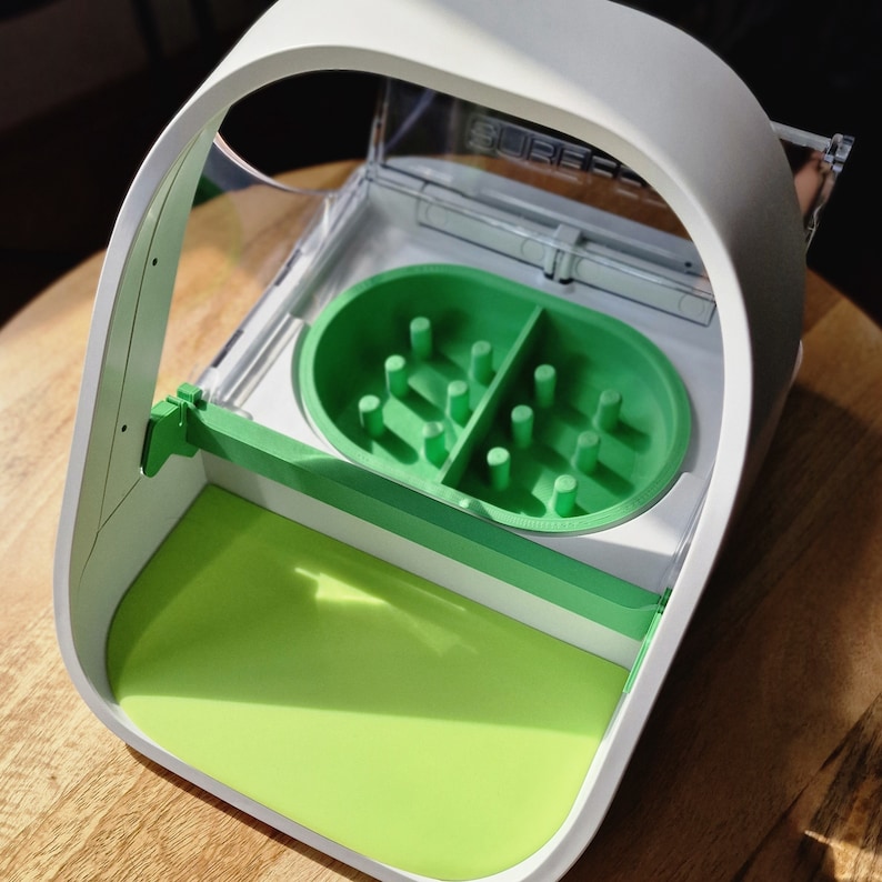 May include: A white plastic maze toy with a green maze and a green lid. The maze has a series of holes and a green ball that can be rolled through the maze.