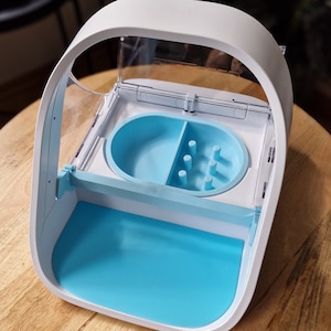 May include: A white plastic device with a clear lid and a blue interior. The device has a small compartment with a blue divider and a section with multiple small holes. The device is designed for storing and dispensing small items.