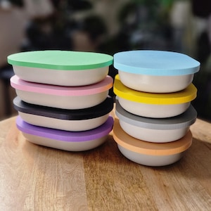 May include: A stack of ten oval-shaped containers with colorful lids. The lids are in the following order from top to bottom: light blue, pink, yellow, gray, purple, orange, black, green, white, and brown.