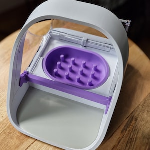 May include: A white and purple pet feeder with a clear plastic lid and a purple dish with multiple holes. The feeder has a white base and a purple handle.