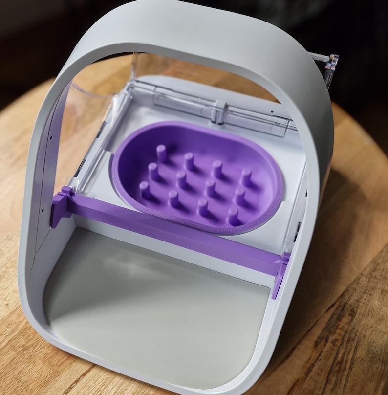 May include: A white and purple pet feeder with a clear plastic lid and a purple dish with multiple holes. The feeder has a white base and a purple handle.