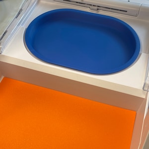 May include: A blue oval-shaped tray sits inside a white compartment. The tray is a vibrant blue color and has rounded edges. Below the tray, an orange surface is visible, creating a contrast in colors. The tray appears to be made of a durable material.