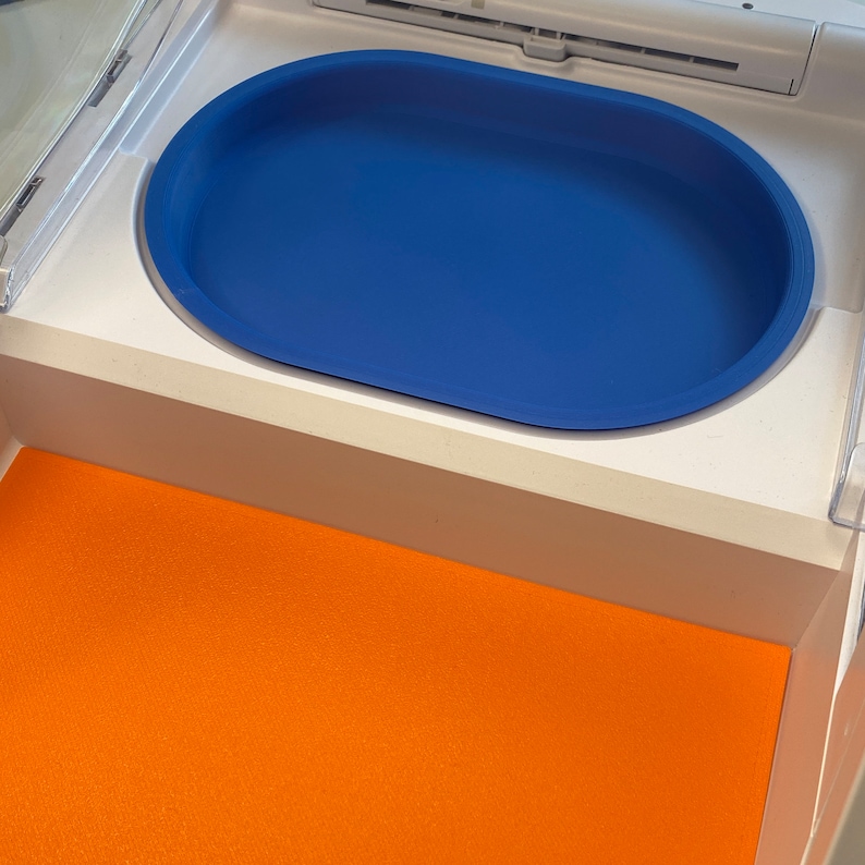 May include: A blue oval-shaped tray sits inside a white compartment. The tray is a vibrant blue color and has rounded edges. Below the tray, an orange surface is visible, creating a contrast in colors. The tray appears to be made of a durable material.
