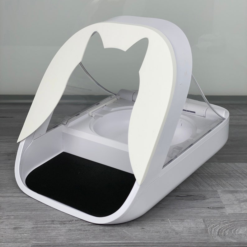 May include: White automatic pet feeder with a clear plastic lid and a black mat. The feeder has a cat-shaped cutout in the front.
