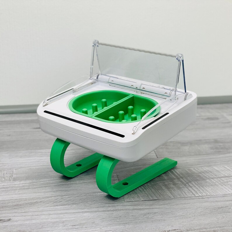 May include: A white and green plastic device with a clear lid. The device has two compartments with green inserts and multiple small holes. The device is on a green stand.