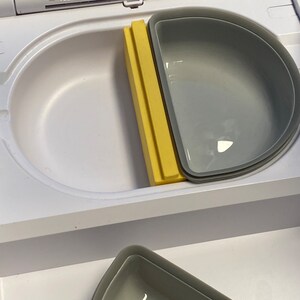 Peut inclure: Un r&eacute;cipient en plastique blanc avec un s&eacute;parateur jaune et deux bols en silicone gris. Le r&eacute;cipient a un couvercle avec une serrure et un bouton.