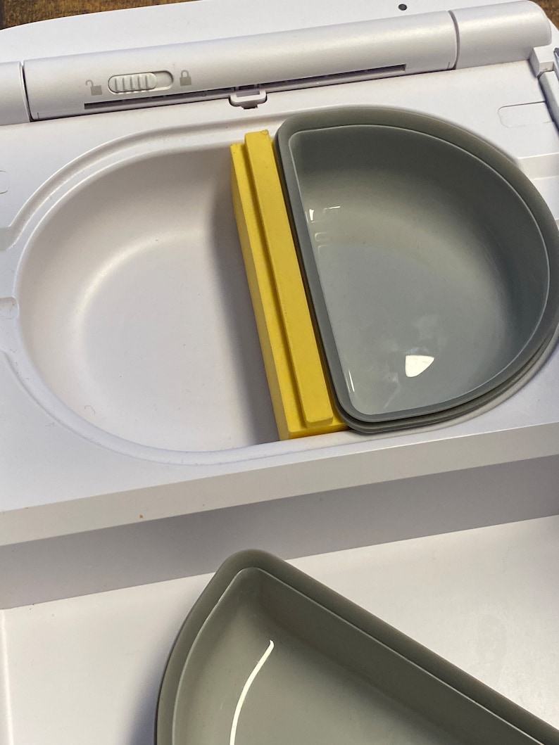 Peut inclure: Un r&eacute;cipient en plastique blanc avec un s&eacute;parateur jaune et deux bols en silicone gris. Le r&eacute;cipient a un couvercle avec une serrure et un bouton.
