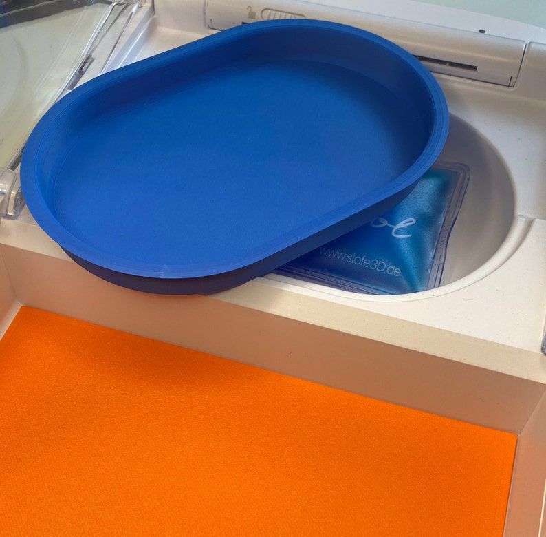 May include: A bright blue, oval-shaped tray sits atop a white compartment. Inside the compartment is a blue gel pack with the text "slofe" and a website address. An orange surface is visible below the tray.