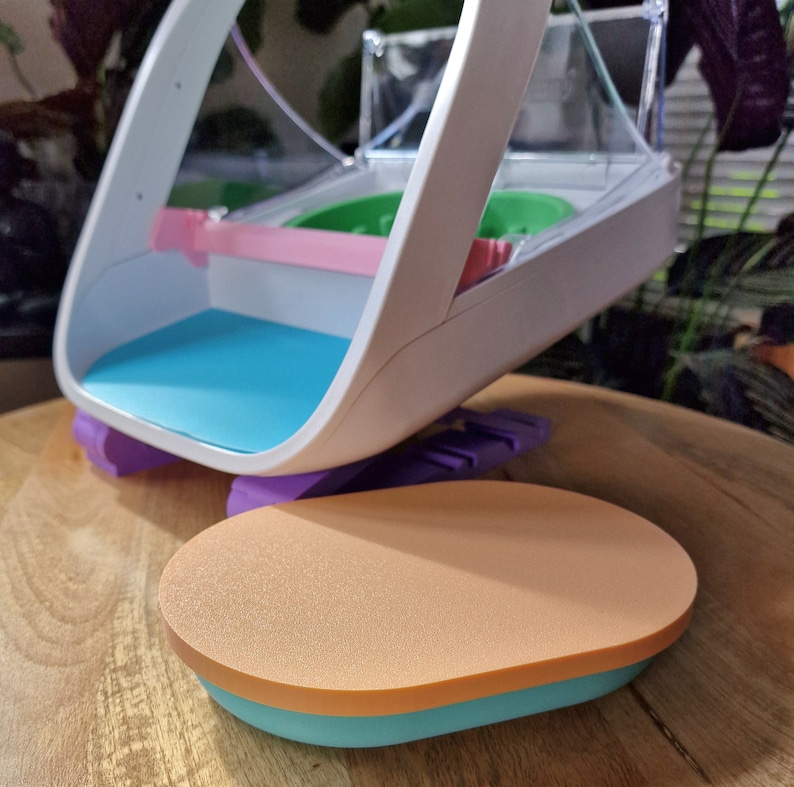 May include: A white plastic toy with a blue and orange base. The toy has a clear plastic top and a pink and green interior. The toy is designed to look like a basketball hoop.