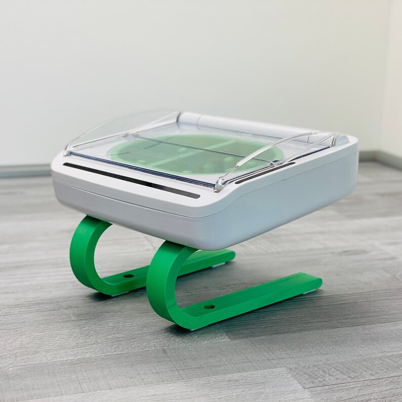 May include: A white, rectangular device with a clear plastic lid sits on a green, C-shaped stand. The device has a green, circular area in the center.