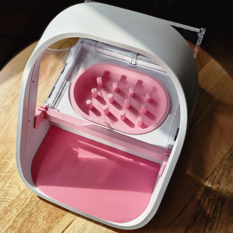 May include: White and pink plastic device with a clear lid and a pink tray with multiple holes. The device is designed for storing and organizing small items.