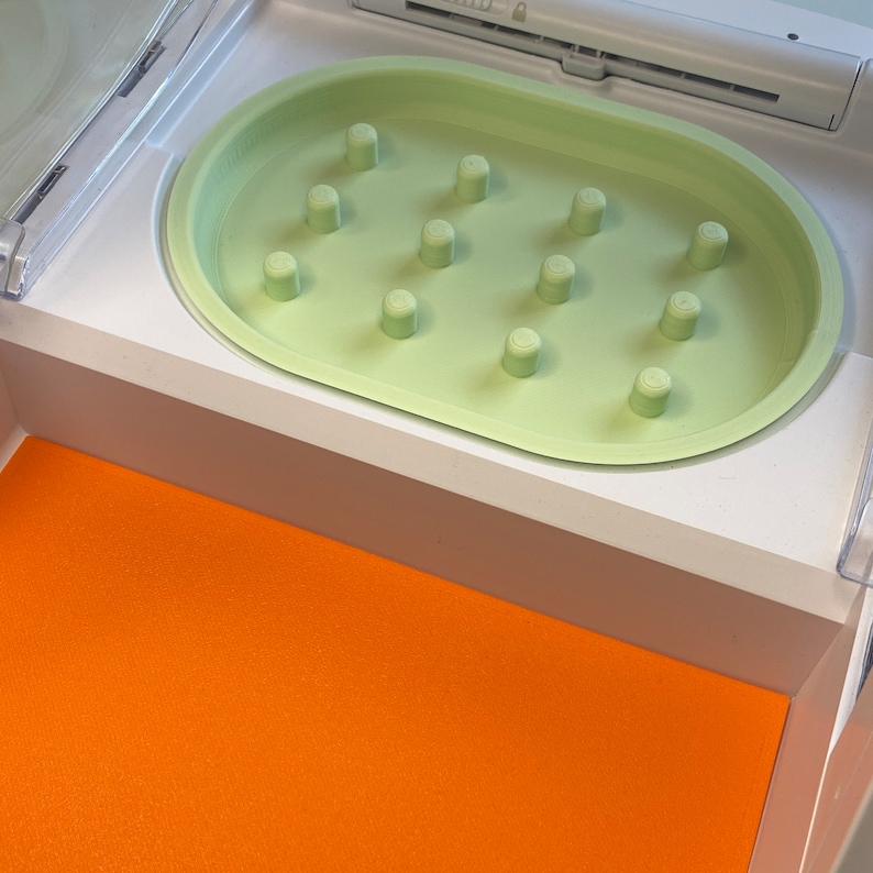 May include: A light green, oval-shaped tray with cylindrical pegs, set within a white compartment. Below, an orange surface provides a contrasting backdrop. The tray appears to be a component of a larger device or organizer.