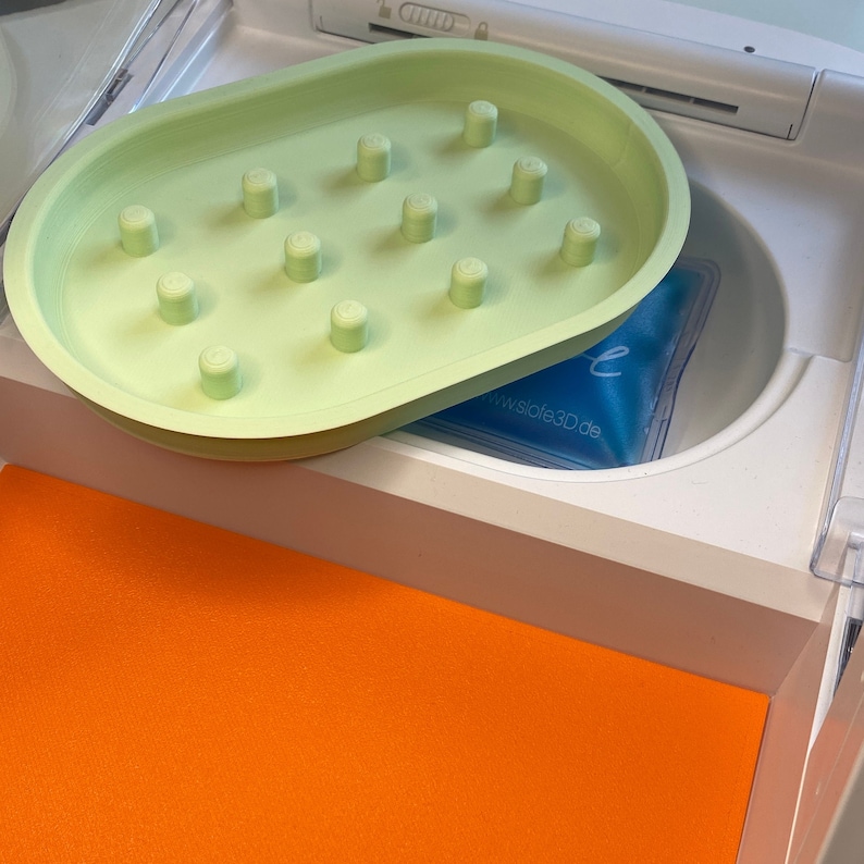 May include: A light green, oval-shaped soap dish with raised cylindrical pegs. The dish sits above a white compartment containing a blue gel pack. An orange surface is visible below the compartment.