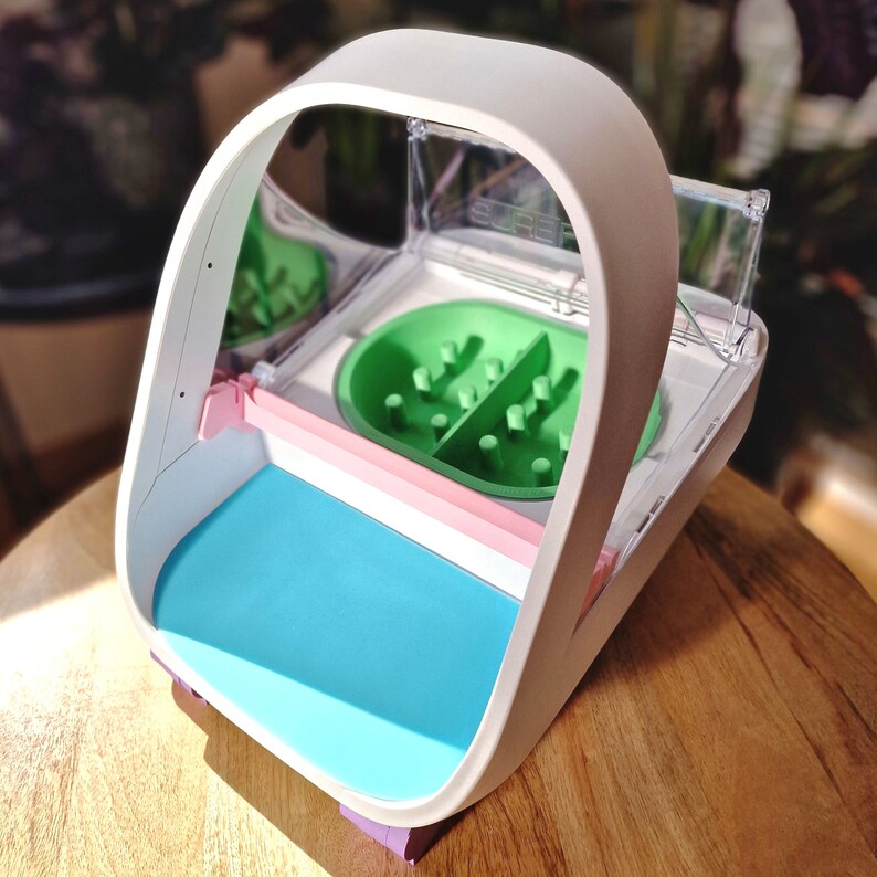 May include: A white plastic interactive pet feeder with a clear plastic lid and a green and pink maze inside. The feeder has a blue base.