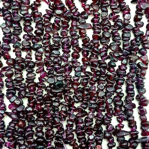 May include: Close-up of strands of dark red-purple gemstone beads. The beads are irregularly shaped and strung together. The beads are a deep, rich colour, with variations in tone. The background is white.