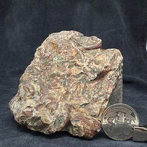 May include: A rough, multi-coloured rock specimen featuring a complex mix of earthy brown, deep red, and muted green tones with a textured, uneven surface. A silver-coloured coin is placed next to the rock to provide a sense of scale for the object.