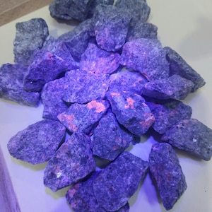 Puede incluir: Un montón de rocas rugosas de color gris oscuro con parches de color naranja brillante y rosa. Las rocas están iluminadas con una luz púrpura, creando un contraste llamativo. Parecen ser especímenes minerales en bruto.