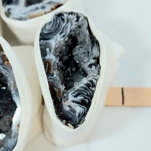 May include: Close-up of a geode slice with a white, gray, and black interior. The geode is encased in a white fabric. The geode's crystalline structure is visible, with bands of color and a hollow center.