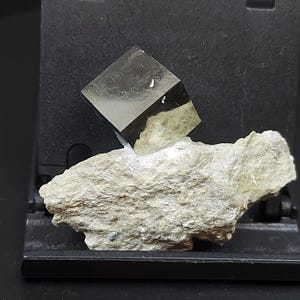 May include: A cubic pyrite crystal with a reflective, metallic surface rests on a rough, beige rock. The pyrite cube is a dark, shiny grey colour, contrasting with the lighter, textured rock base. The composition is set against a dark background.