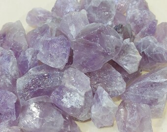 May include: A pile of raw amethyst crystals in varying sizes and shapes. The crystals display a range of purple hues, from pale lavender to deep violet. The rough, natural texture of the stones is visible, with some having sharp edges and others appearing more rounded.