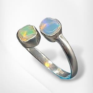 May include: A silver ring with two opal gemstones. One opal has green and yellow colours, the other blue and white. The ring's band splits, each end holding an opal. The ring is made of silver.