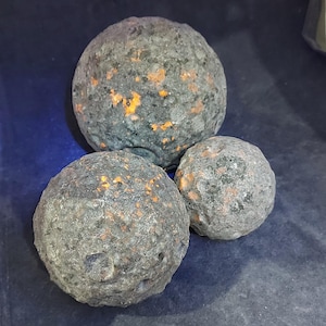 May include: Three spherical, grey stones with orange and yellow speckles. The stones have a rough, textured surface and vary in size, with the largest stone at the top and two smaller stones below.