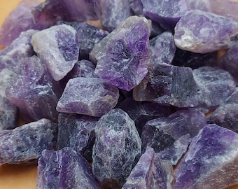 May include: A pile of raw amethyst crystals in varying shades of purple. The rough-cut stones have a natural, unpolished appearance, showcasing their crystalline structure. The crystals are a mix of light and dark purple hues.