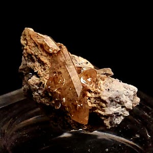 Op de afbeelding: Close-up van een mineraal specimen met meerdere, doorschijnende, amberkleurige kristalformaties. De kristallen zijn geplaatst op een ruwe, beige matrix. De kristallen hebben scherpe randen en een glasachtig uiterlijk. Donkere achtergrond.