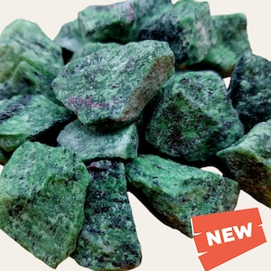 May include: A pile of rough zultanite gemstones in green and red hues. The stones are irregularly shaped and have a mottled appearance.