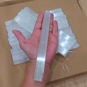 Puede incluir: Una mano sostiene una varita larga de selenita blanca translúcida, con varias varitas similares y piezas de selenita en el fondo. Las varitas tienen un aspecto liso y pulido, y las piezas de selenita están apiladas sobre una superficie marrón.