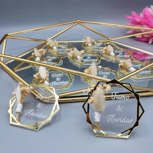 Könnte beinhalten: Ein Set aus 10 goldgerahmten, transparenten Acryl-Platzkarte mit den eingravierten Namen "Jamo & Hevidar". Jede Karte ist mit einem kleinen Strauß getrockneter weißer Blumen verziert.