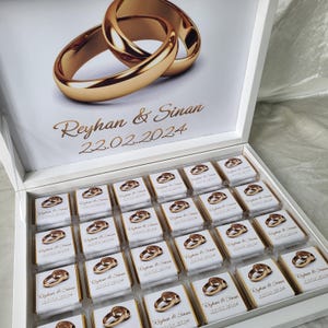 Op de afbeelding: Een witte doos met een deksel, open om een ​​lade met kleine witte doosjes te onthullen met gouden trouwringen erop gedrukt. Het deksel heeft een gouden ringontwerp en de tekst "Reyhan & Sinan 22.02.2024".