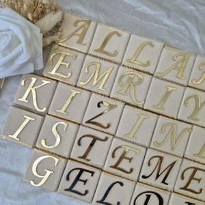 Op de afbeelding: Een set gouden letters op een beige stoffen achtergrond. De letters spellen de woorden "ALEMRIY KIZSTEME" uit.
